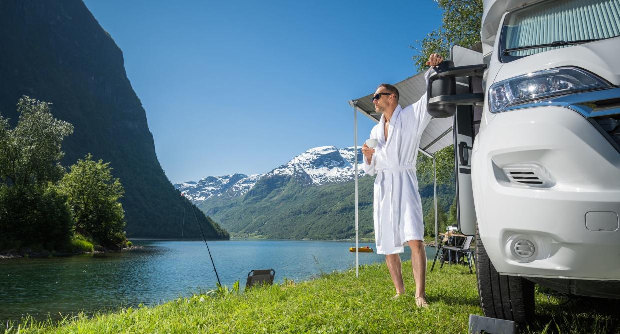 Tourist in bath robe with a cup of coffee in front of RV and picturesque mountain lake scene