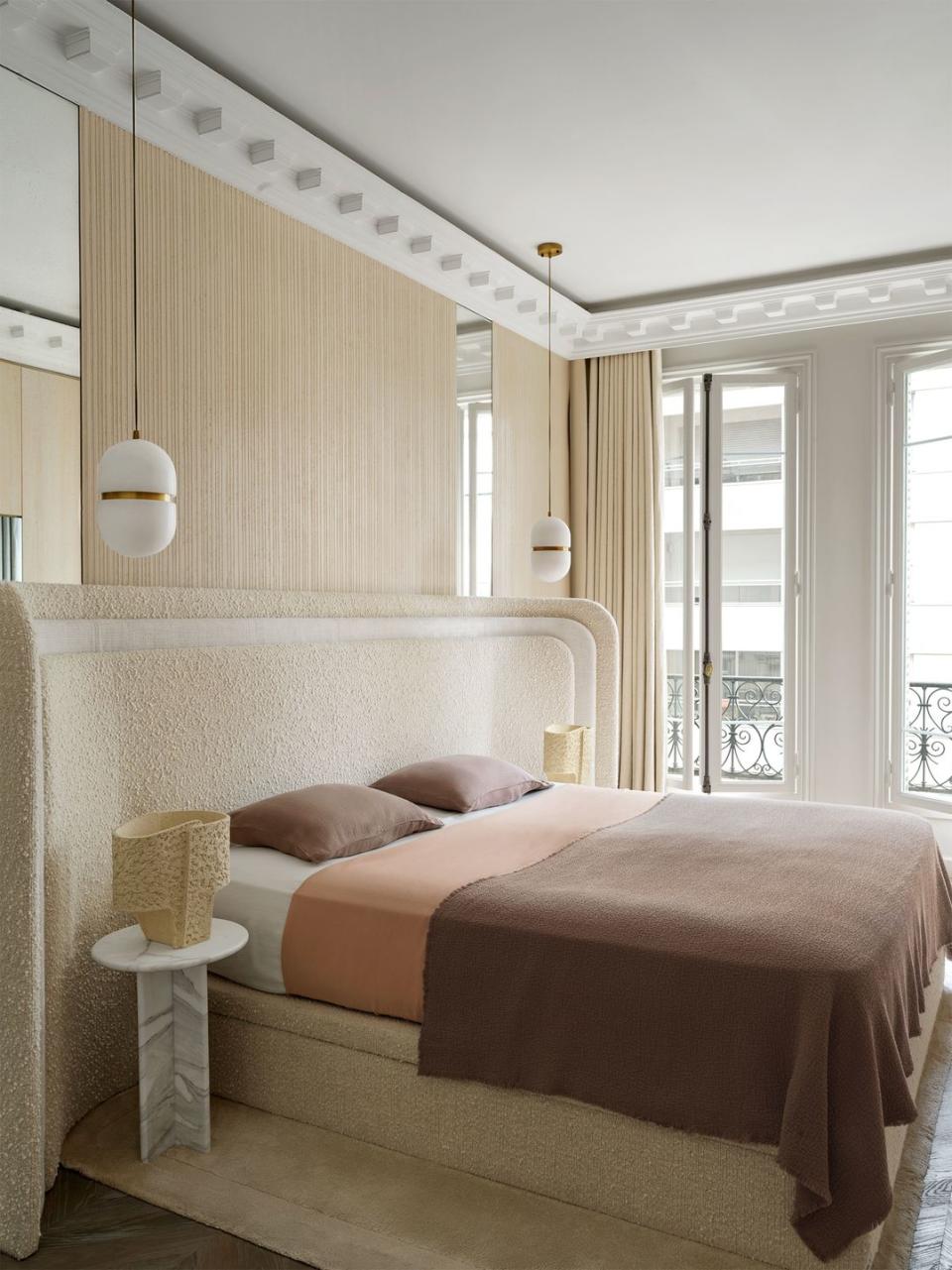 a bed with upholstered curved headboard and frame with brown and peach bedding, marble nightstands with lamps, grooved wall treatment, suspended pendants, curtained windows to a small terrace