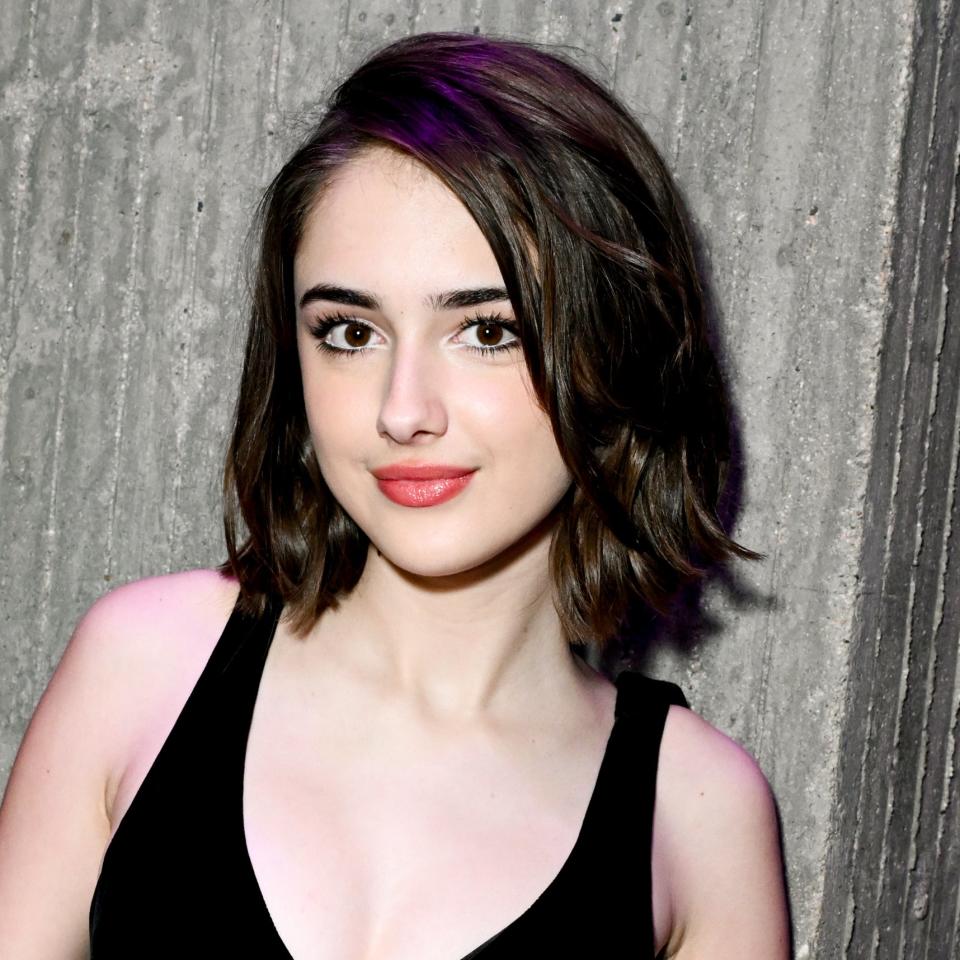 Julia Butters with shoulder-length hair in a black dress and scarf stands in front of a textured wall and smiles at the camera