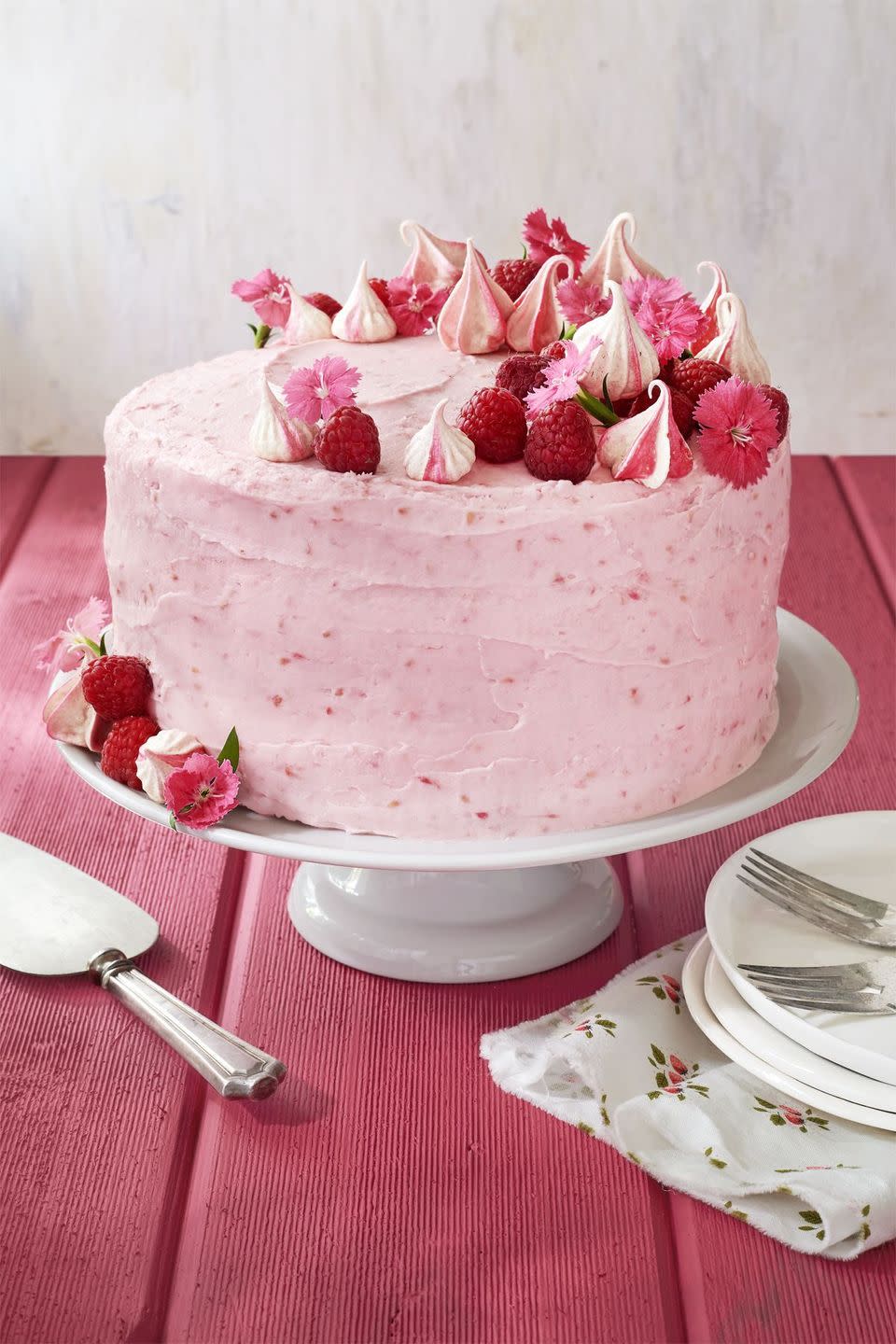 raspberry pink velvet cake with raspberry cream cheese frosting on a white cake stand and garnished with fresh raspberries small pink flowers and pink swirled meringue kisses