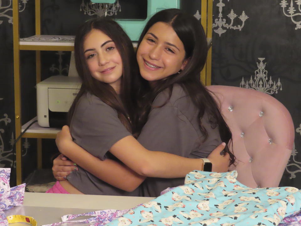 Audrina Demma, left, and her sister Giuliana hug in the basement at the sewing station of their Freehold, N.J., home on June 19, 2024. The sisters have sewn and donated 1,800 hospital gowns to sick children in 36 states plus Africa. Giuliana was inspired by seeing her baby cousin wearing an ugly, ill-fitting gown while hospitalized with brain cancer in 2017. (AP Photo/Wayne Parry)