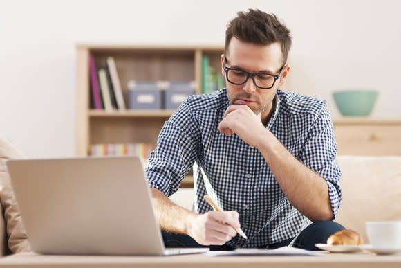 A man looking at material on his open laptop and making notes on paper.