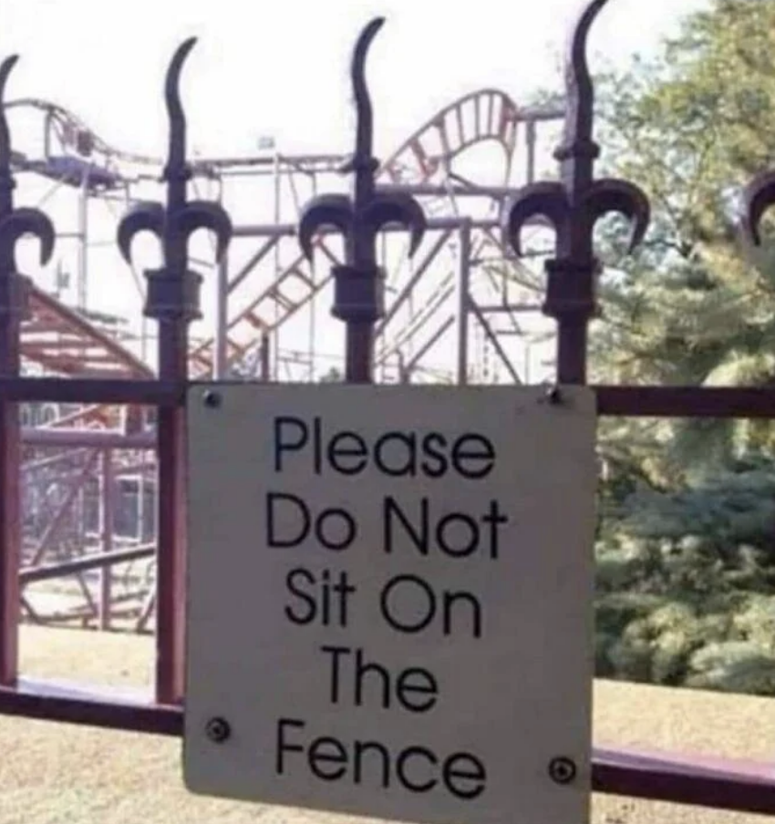 Sign on a fence that reads, "Please Do Not Sit On The Fence," with a roller coaster visible in the background