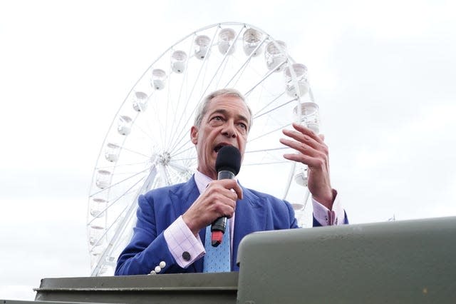 Nigel Farage speaks into a microphone on a grey day with a large Ferris wheel behind him