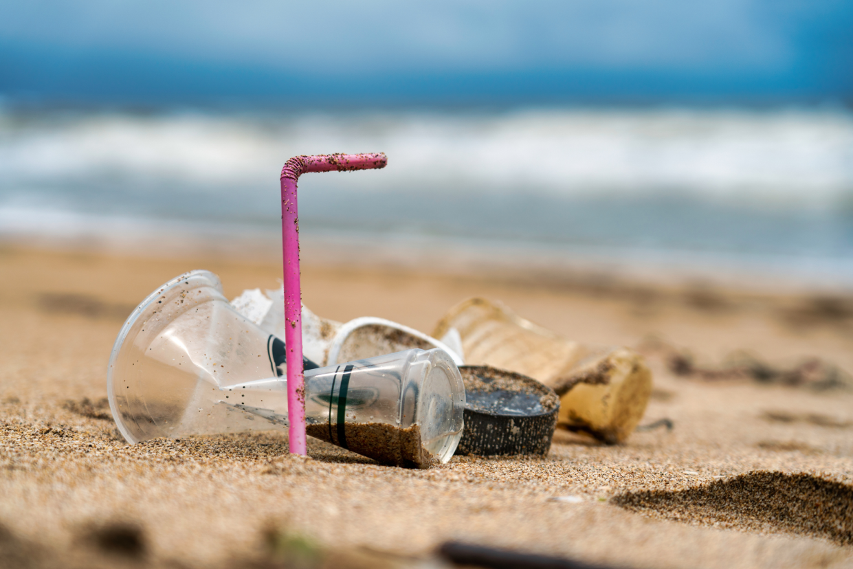 Selective focus on one pink plastic straw in 'L' shape standing in the sand with three plastic coffee cups with ocean in the background