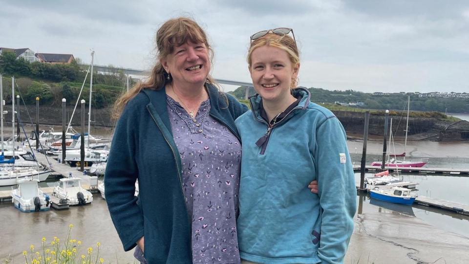 A mum and daughter stand in front of a marina with their arms around each other smiling at the camera