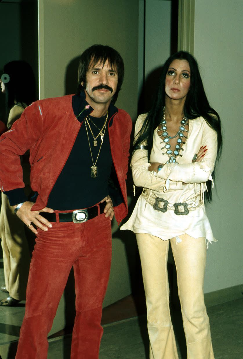 sonny bono and cher stand next to each other inside, he wears a matching red jacket and pants with a black shirt and several necklaces, she wears a white top with yellow pants and several necklaces