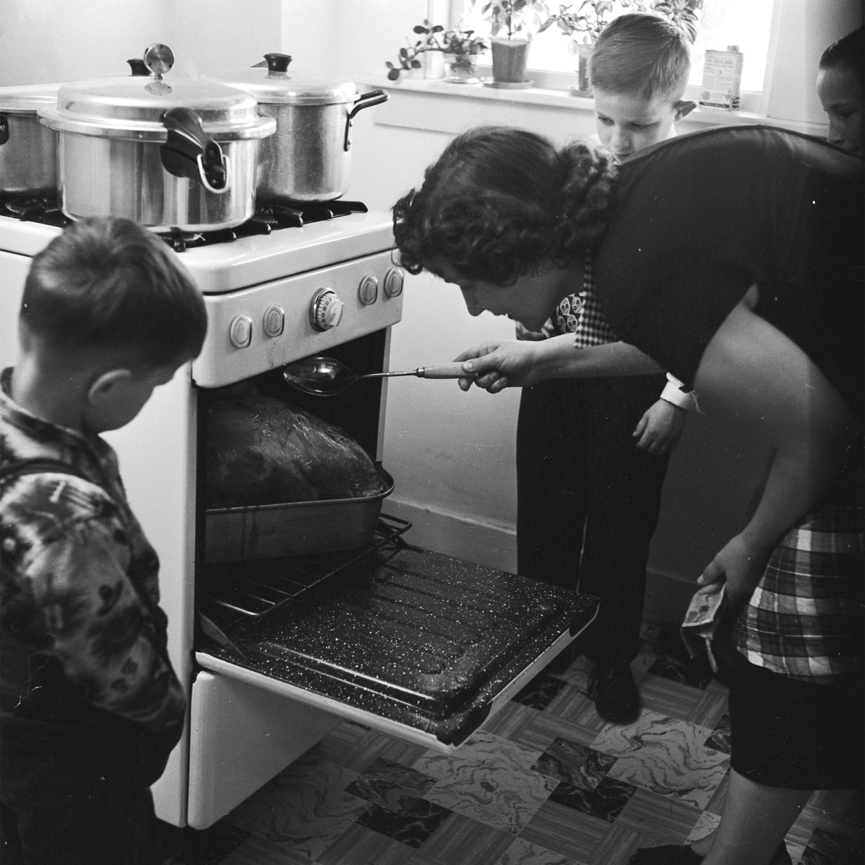 An American housewife, Mrs Boivin, bastes the Thanksgiving turkey while her two sons look on.