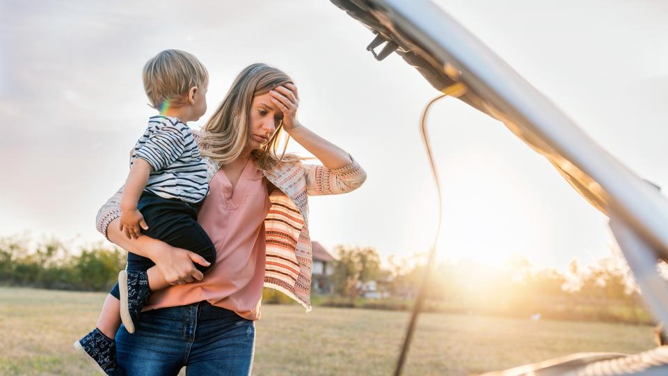 Portrait of young Caucasian woman standing by broken down car and waiting for assistance while holding her baby boy during sunset.