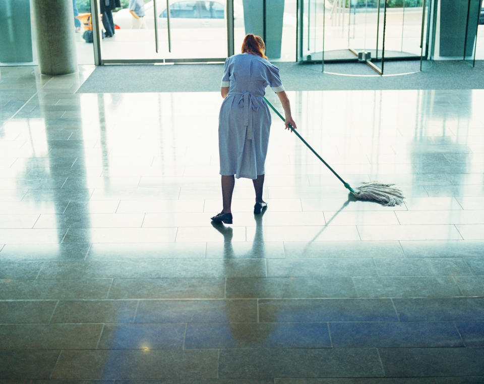 A janitor in uniform cleans a large tile floor in the lobby of an office building, with sliding doors and windows in the background