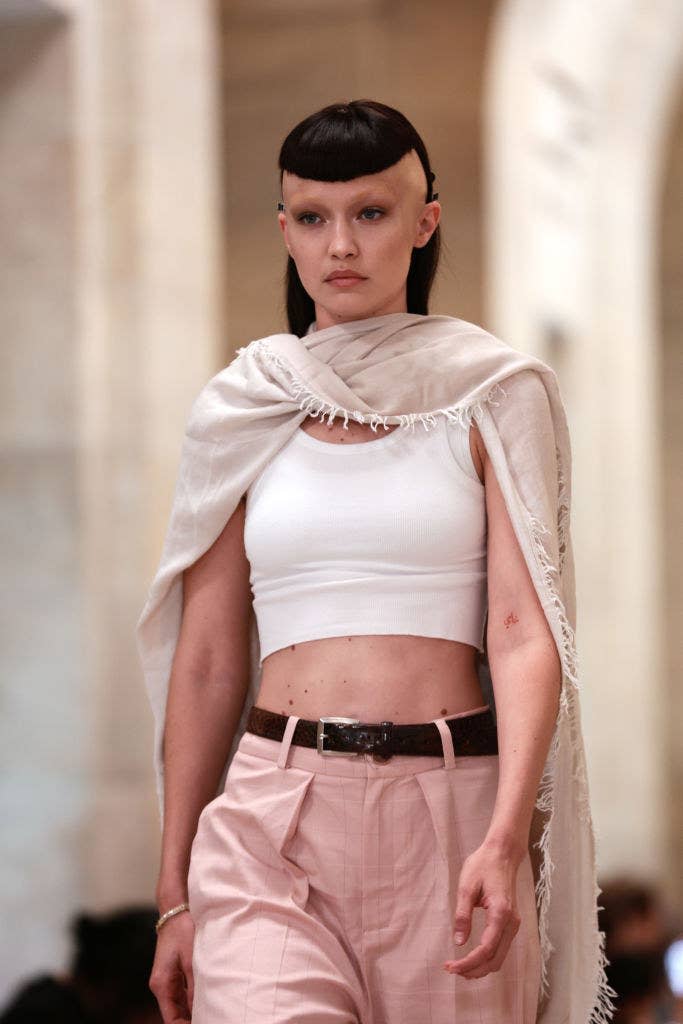 One of the sisters with a bowl haircut with the sides of her head shaved, wearing a crop top and shawl