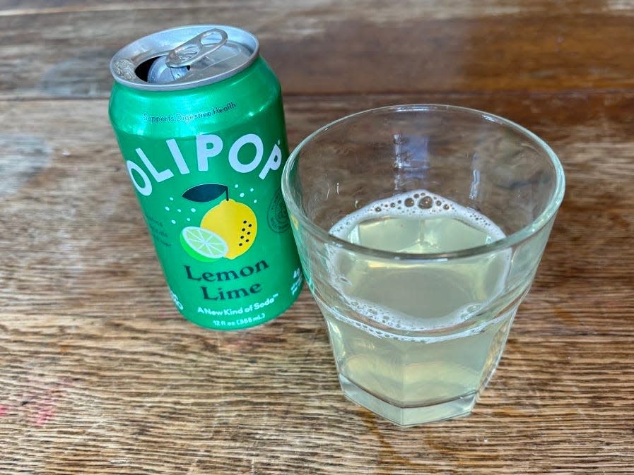 An open can of lemon-lime Olipop next to a small, clear glass with pale-yellow liquid inside. Both are sitting on a wooden table.