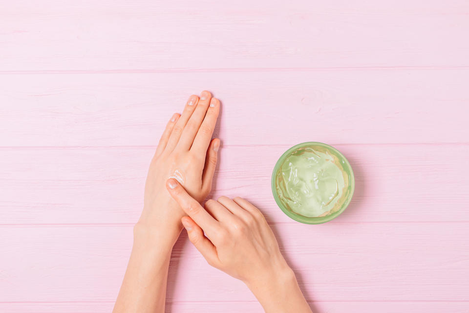 Female hands apply aloe gel on skin next to jar of cosmetic product on pink table, flat lay.