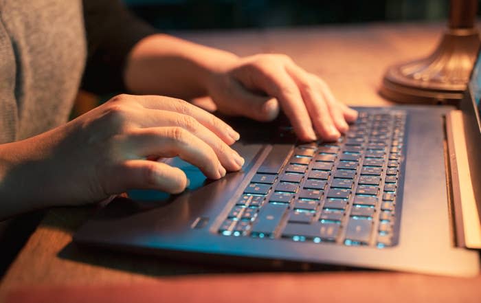 Hands typing on a laptop keyboard at a wooden desk, illuminated by a nearby lamp