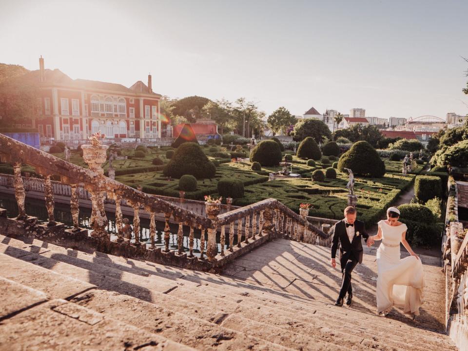 A groom helps a bride walk up a set of stairs next to an extravagant garden and home.