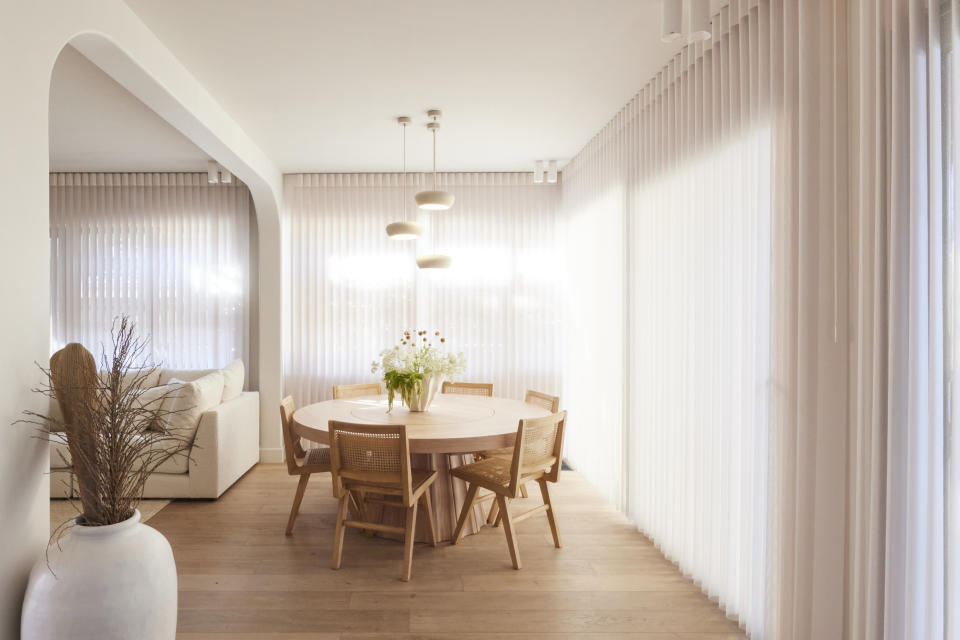 A wide shot of the dining room, with the table in the centre a large potted plant on the left and white sheer curtains on the back and right wall.