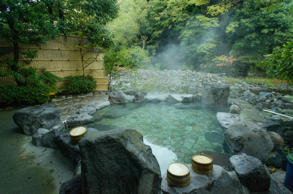 Tranquil outdoor hot spring with steam rising, surrounded by rocks and greenery, and wooden buckets by the side
