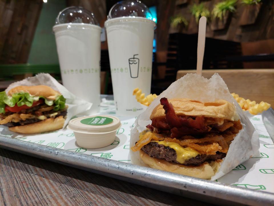 A selection of food from Shake Shack, displayed on a tray in a restaurant: a crispy shallot burger, a ShackMeister with bacon, Shack sauce, cheesy fries, two drinks