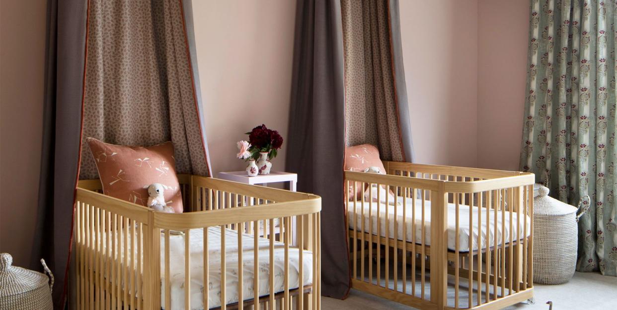 twin cribs with matching valanced canopies in a nice brown against a dusky pink wall and on the whitish carpet is a series of small animal figures in a row and the ceiling is covered in a wavy yellow orange and white pattern with a large leafy off white chandelier at center