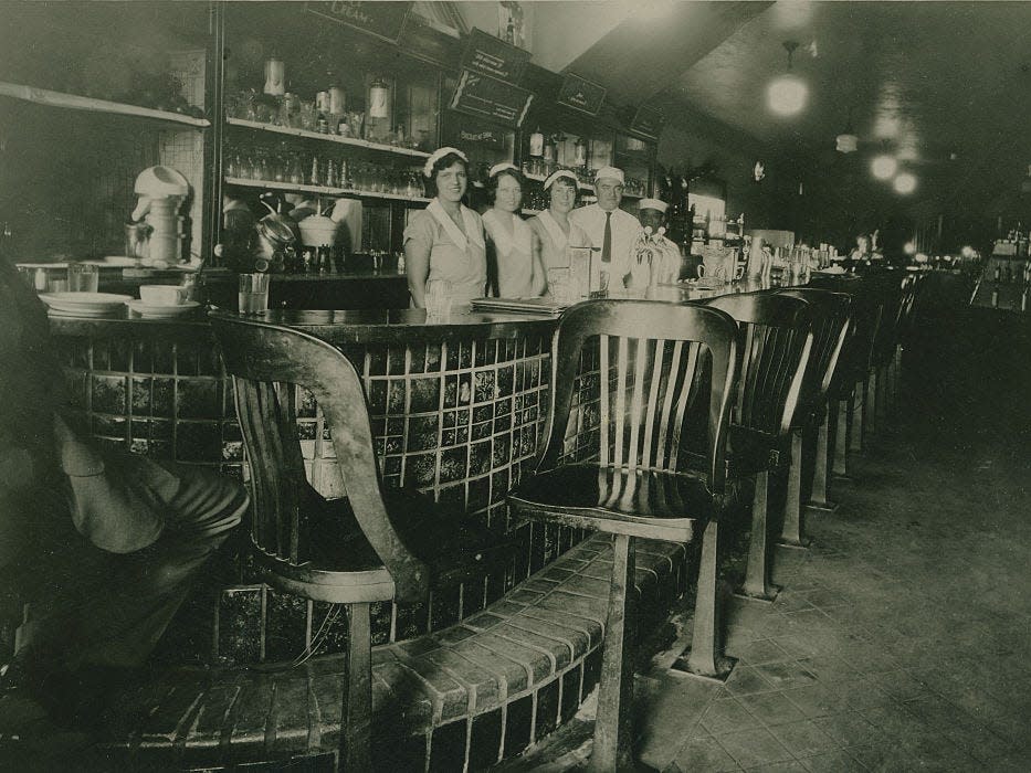 Black and white photograph of staff in uniform posed standing behind bar in 1924