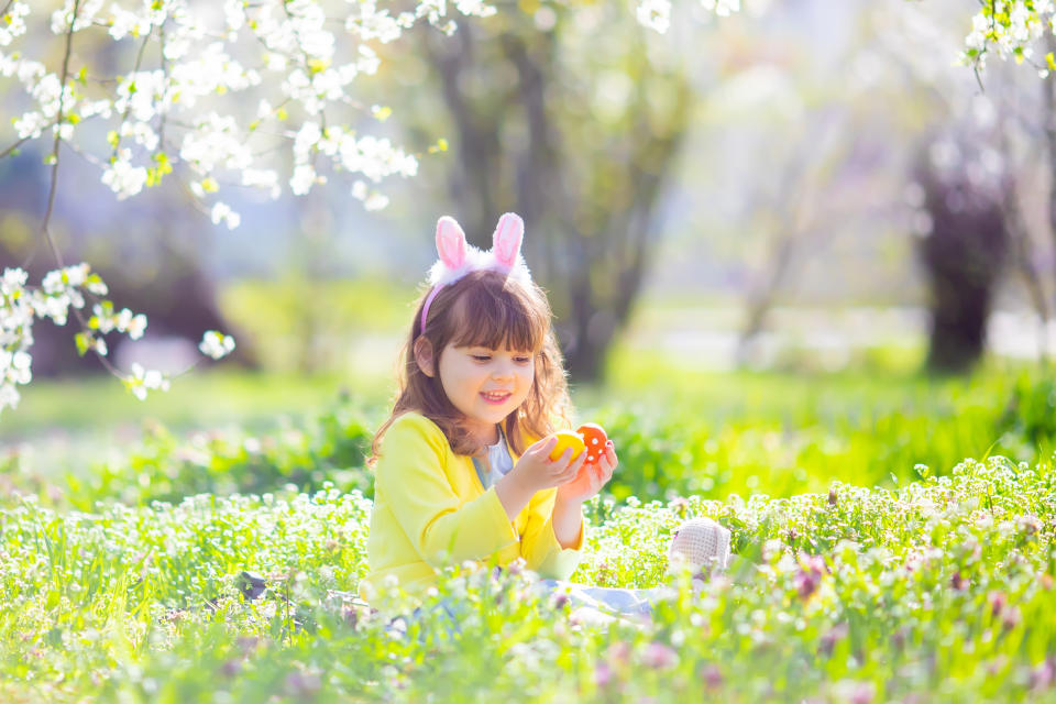 Cute little girl with curly hair wearing bunny ears and summer dress having fun during Easter egg hunt relaxing sitting at the grass and playing with colorful easter eggs in the blooming garden on a sunny spring day