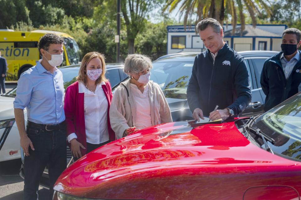 Gov. Gavin Newsom signs an executive order on the hood of a red car outdoors while four people look on.