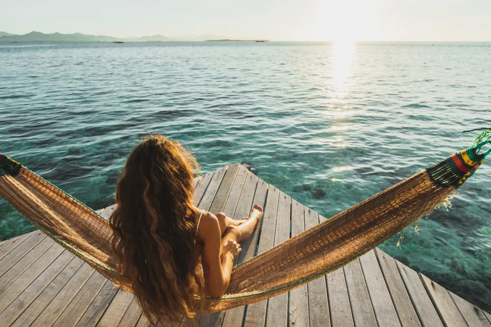 A woman with long hair relaxes in a hammock on a wooden pier, overlooking a serene ocean at sunset