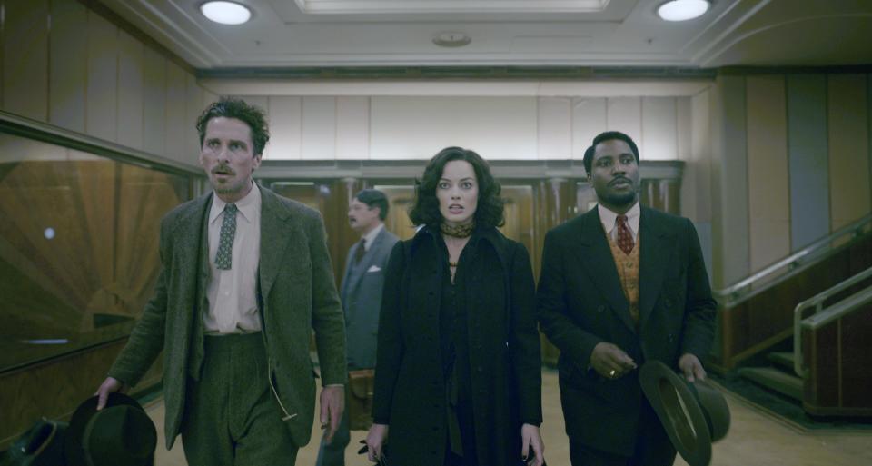 Christian Bale, Margot Robbie, and John David Washington in vintage attire, walking through an Art Deco-inspired hallway, looking ahead with serious expressions