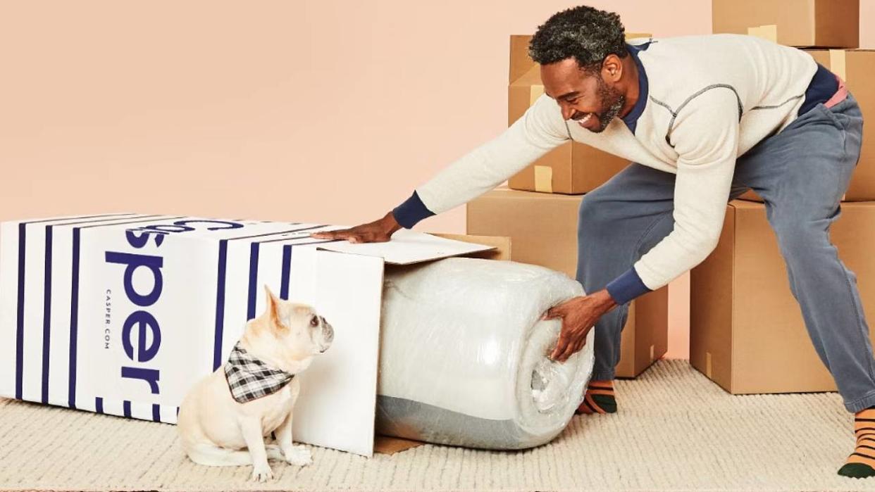  A man pulling a bed in a box mattress out of a Casper box with a dog sat next to it. 