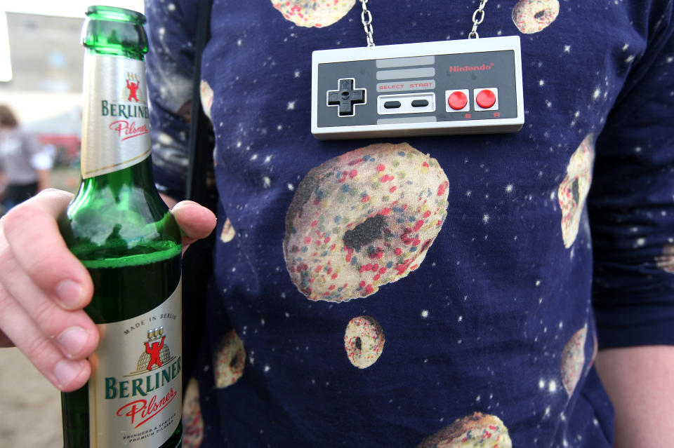 BERLIN, GERMANY - JULY 21: An attendee wearing a 1980s Nintendo controller as a necklace holds a Berliner Pilsener beer at the second annual Hipster Olympics on July 21, 2012 in Berlin, Germany. With events such as the "Horn-Rimmed Glasses Throw," "Skinny Jeans Tug-O-War," "Vinyl Record Spinning Contest" and "Cloth Tote Sack Race," the Hipster Olympics both mocks and celebrates the Hipster subculture, which some critics claim could never be accurately defined and others that it never existed in the first place. The imprecise nature of determining what makes one a member means that the symptomatic elements of adherants to the group vary in each country, but the archetype of the version in Berlin, one of the more popular locations for those following its lifestyle, along with London and Brooklyn, includes a penchant for canvas tote bags, the carbonated yerba mate drink Club Mate, analogue film cameras, asymmetrical haircuts, 80s neon fashion, and, allegedly, a heavy dose of irony. To some in Berlin, members of the hipster "movement" have replaced a former unwanted identity in gentrifying neighborhoods, the Yuppie, for targets of criticism, as landlords raise rents in the areas to which they relocate, particularly the up-and-coming neighborhood of Neukoelln. (Photo by Adam Berry/Getty Images)