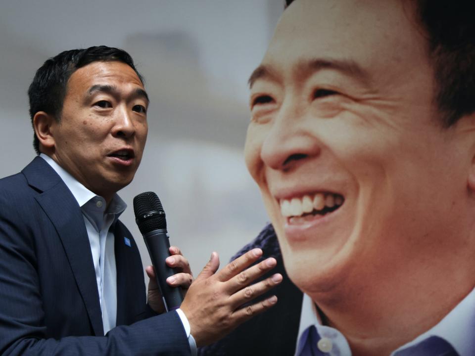 Andrew Yang speaks with a microphone in front of a blown up portrait of his face.