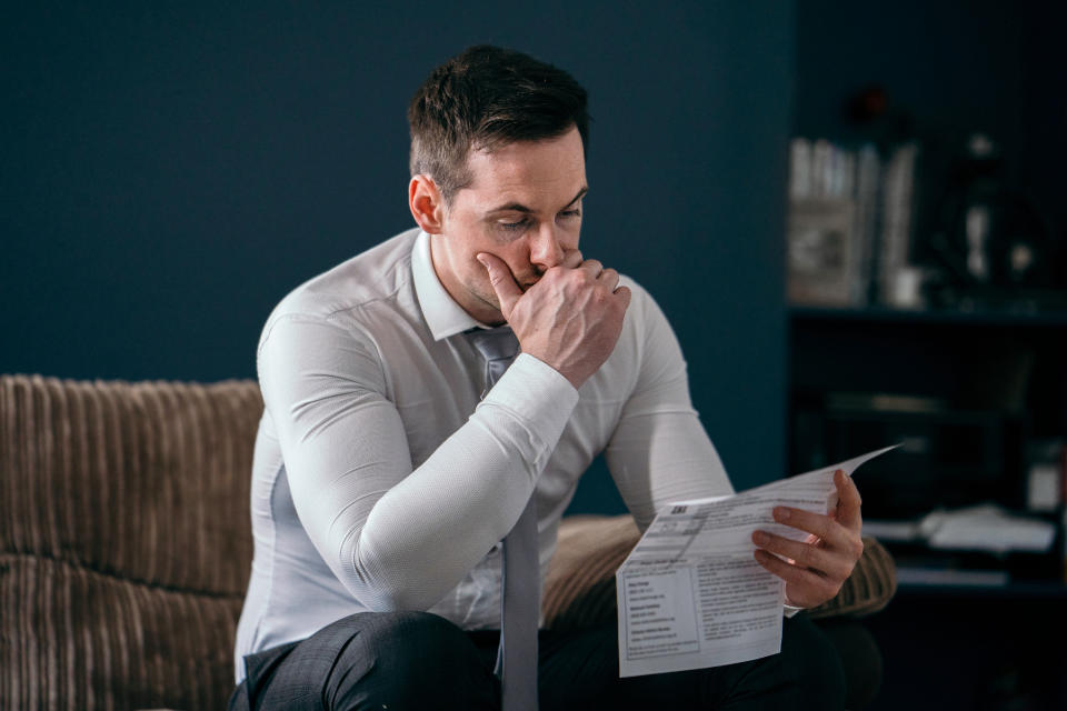 Middle aged man worried about the rise in the energy price cap . Expensive energy bills are causing him stress . He is reading a bill on the sofa of his living room at home . Debt , loans and money saving issues are on his mind.