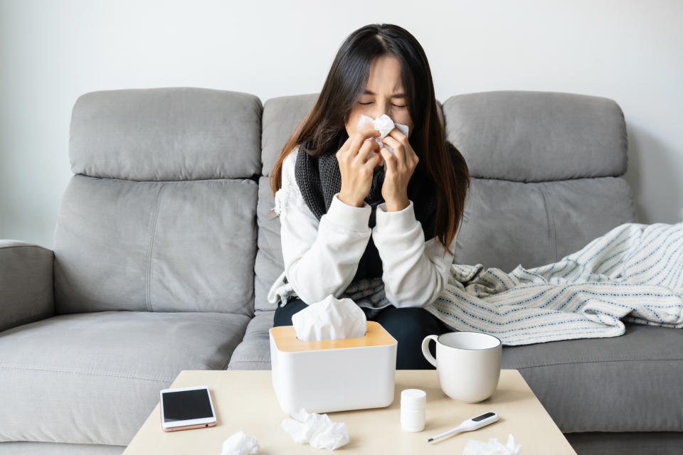 Young Asian woman get sick with changing seasons sitting on couch. Girl feel cold and has a high fever blowing nose while sit on sofa in living room at home. Health problems, allergy concept.