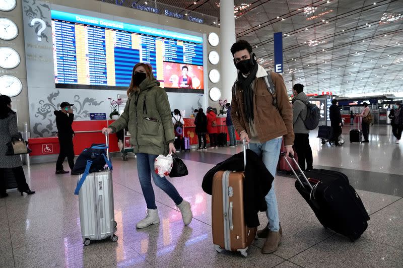 Foreign travellers wearing masks walk past a departures information board at Beijing International Airport in Beijing