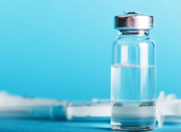 A bottle containing a vaccine and a syringe in the background.