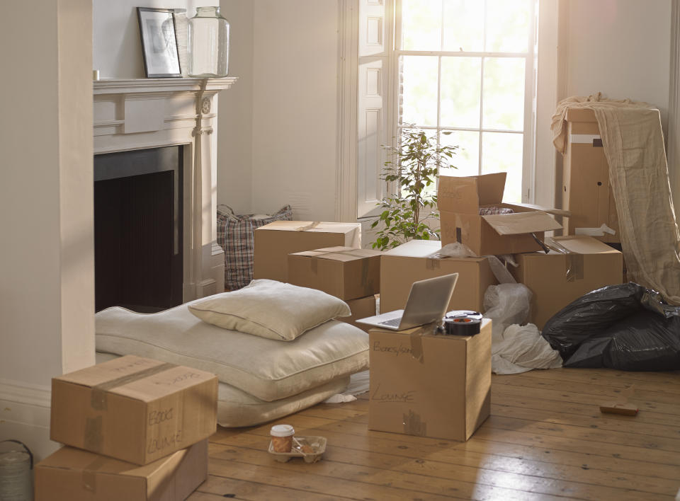 A room filled with unpacked moving boxes, cushions on the floor, a laptop on a box, and a window letting in sunlight