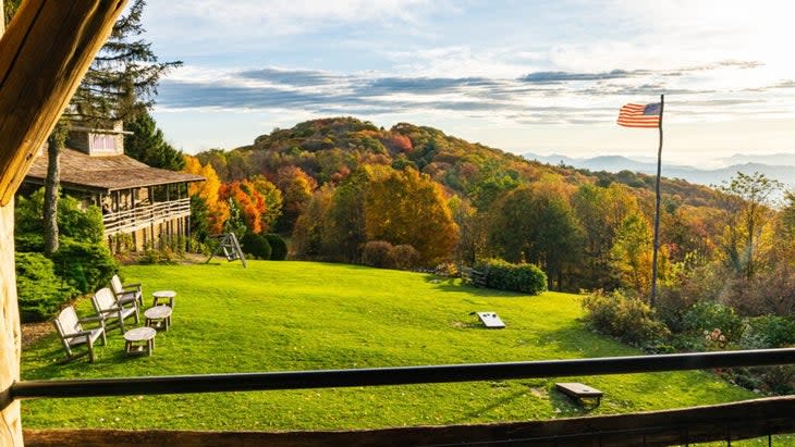 A hillside behind then property covered in brilliant fall foliage. An American flag waving on the side of the property's lush lawn.
