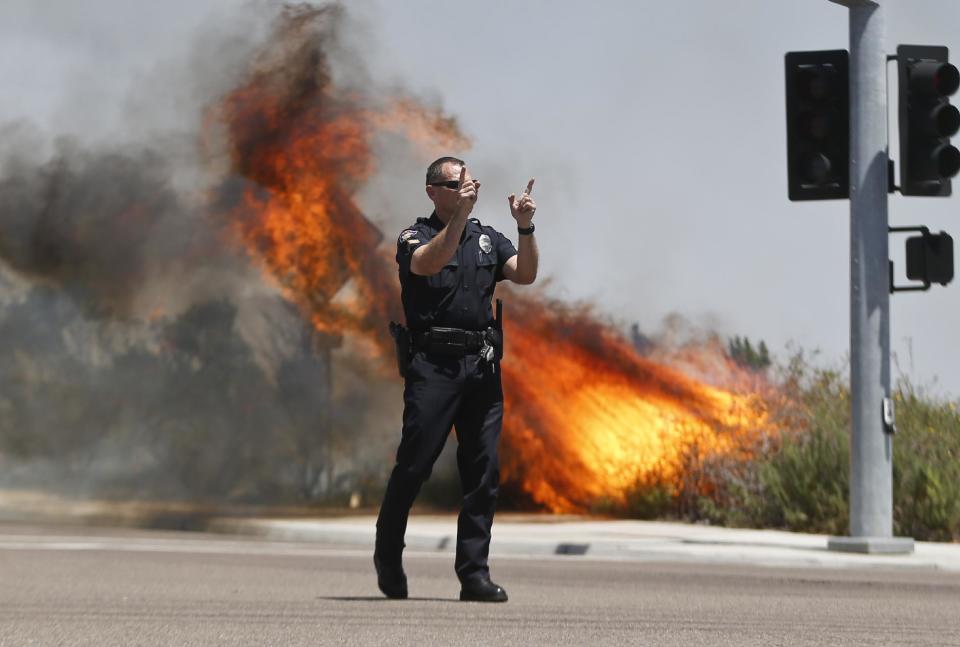 A Carlsbad,Calif. police officer turns traffic away as flames leap behind him Wednesday, May 14, 2014, in Carlsbad, Calif. Weather conditions that at least temporarily calmed allowed firefighters to gain ground early Wednesday on a pair of wildfires that forced thousands of residents to leave their homes. (AP Photo)