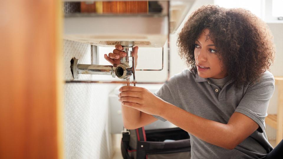 Young black female plumber sitting on the floor fixing a bathroom sink, seen from doorway.