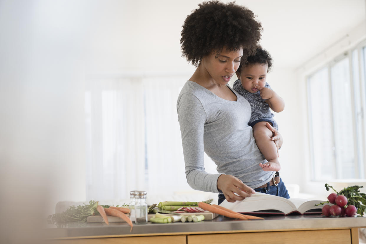 A mother holds her child as she reads a cookbook while prepping veggies, illustrating that how moms can be susceptible to overuse injuries because of repetitive movements like carrying children, according to experts. 