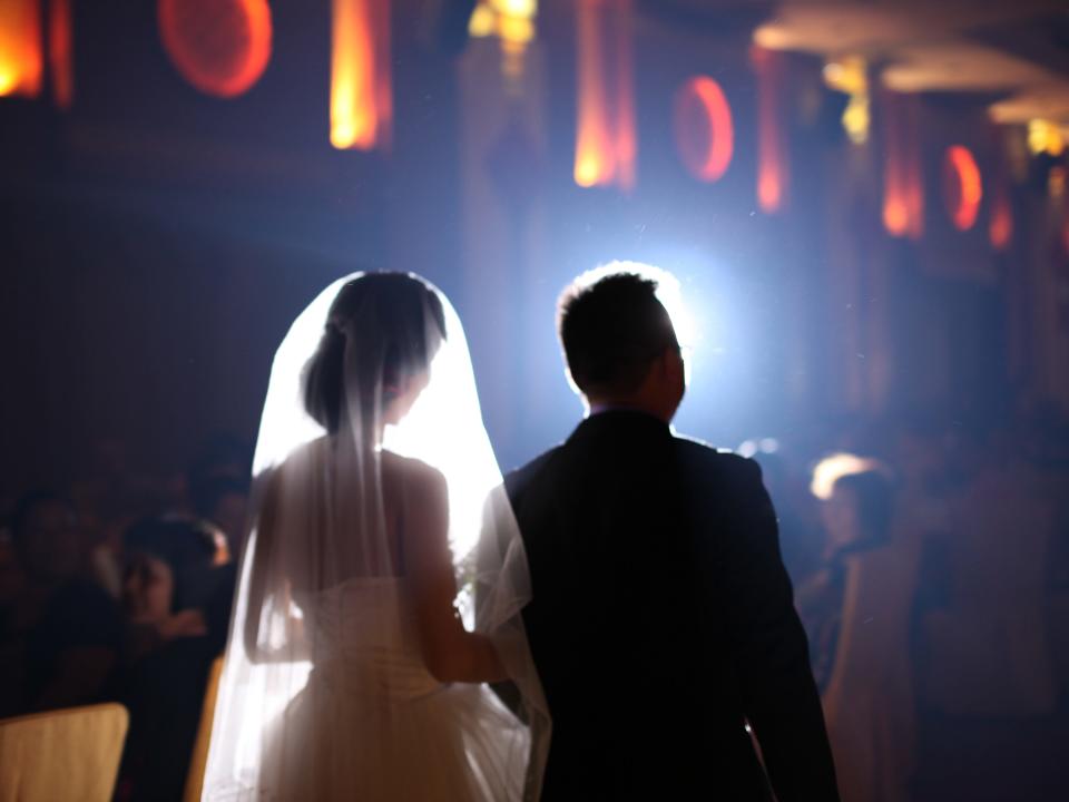 A woman in a wedding dress and a man in a suit walkaway with backs toward camera in dimly-lit room with orange lights along wall