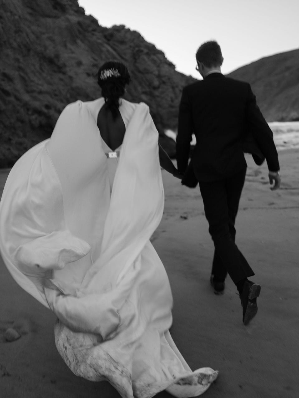 A bride and groom run together in their wedding attire on a beach.