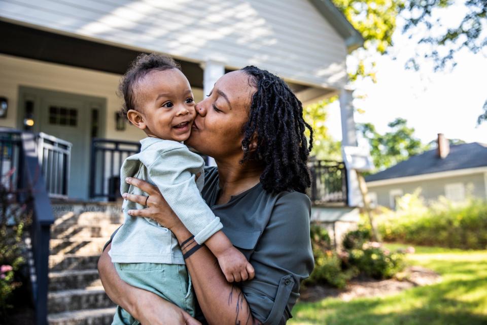 A woman holds a young child outside and kisses their cheek.