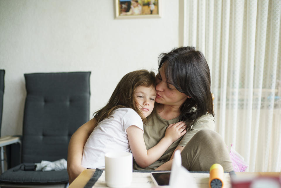 A woman embraces a young girl who looks slightly sad, sitting inside a cozy home. A small picture frame is visible on the wall behind them