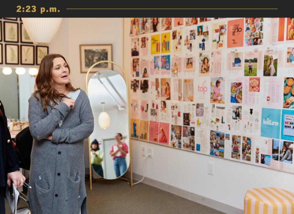 Drew Barrymore, wearing a long gray cardigan, stands in front of a wall covered in magazine pages.