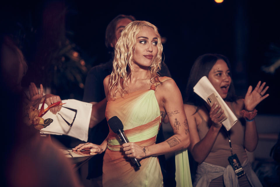 Miley Cyrus, in a stylish one-shoulder dress, holds a microphone while engaging with fans. One fan excitedly reaches out, holding a phone and papers