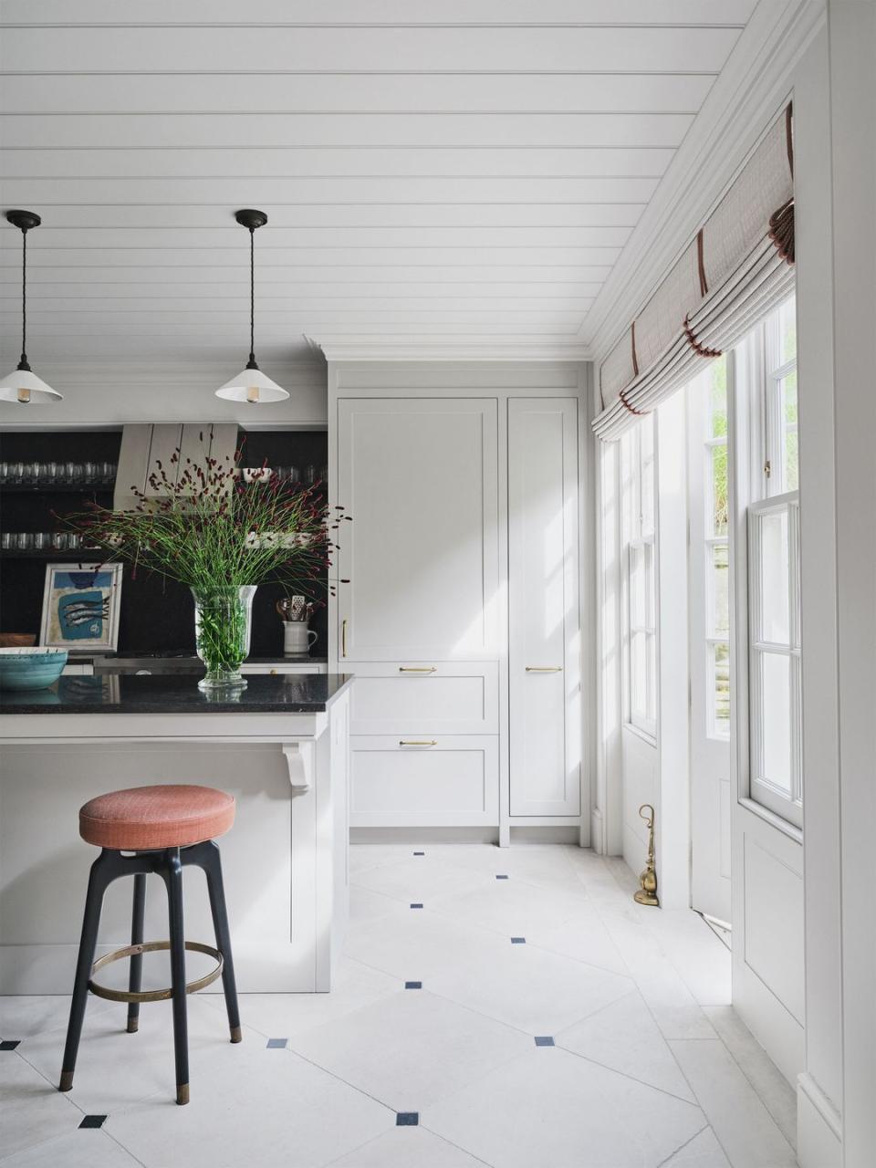 a primarily white kitchen has built in cabinets, windows and a door with rolled shades, glass pendants over an island with a black marble top matching wall behind counter, a stool with fabric seat