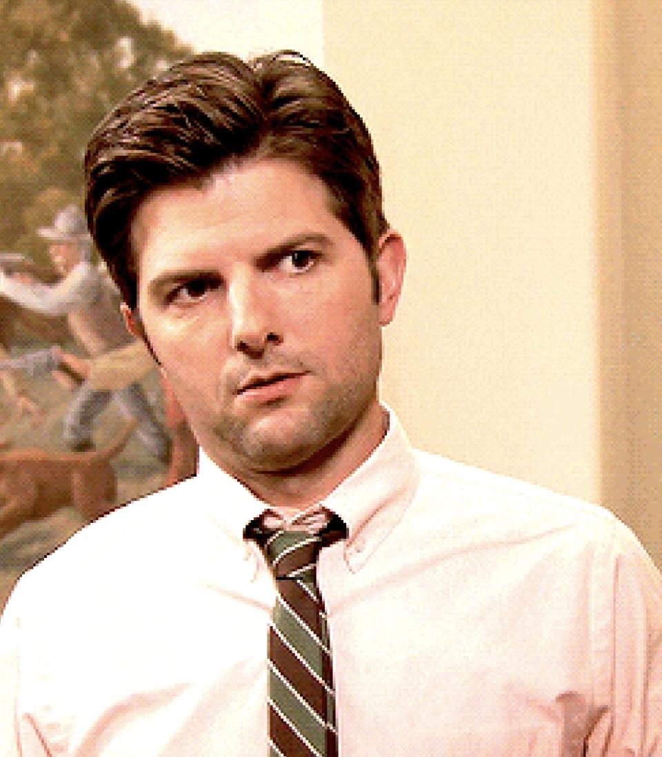 Adam Scott in a scene from a TV show, wearing a dress shirt and striped tie, looking slightly to the side with a neutral expression