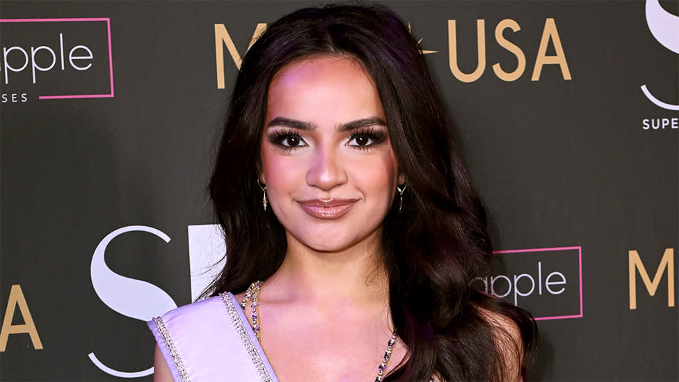 Miss Teen USA 2023, UmaSofia Srivastava smiling at the camera, with a wall behind her which contains names of companies and Miss USA.