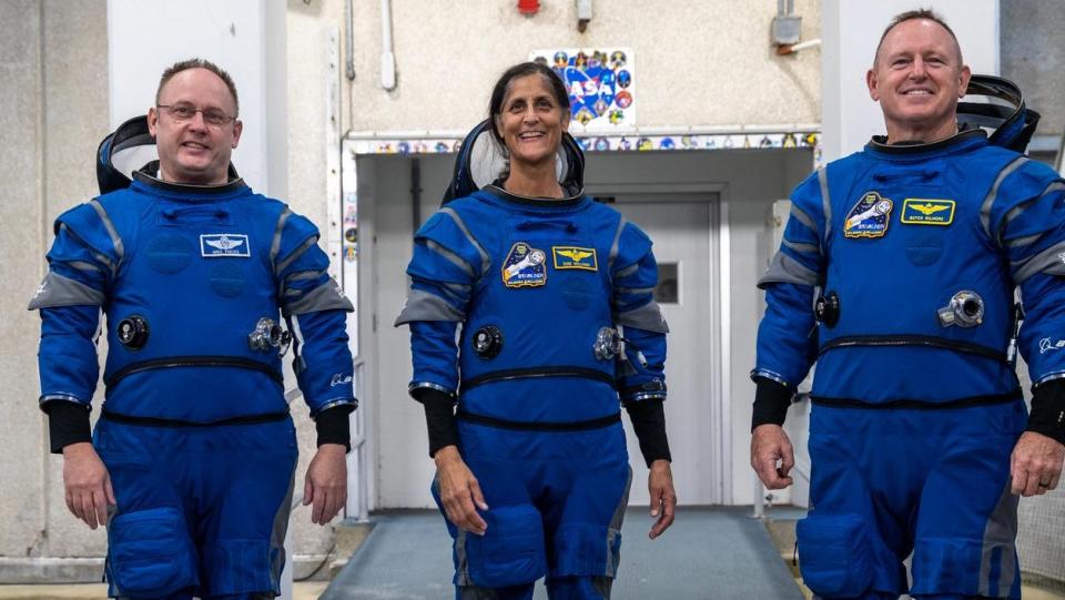 Three astronauts in spacesuits smiling in front of a door with the NASA logo